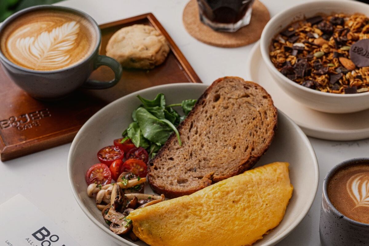A Boojee brunch spread of sourdough toast, an omelette, granola and two coffees on a pale table
